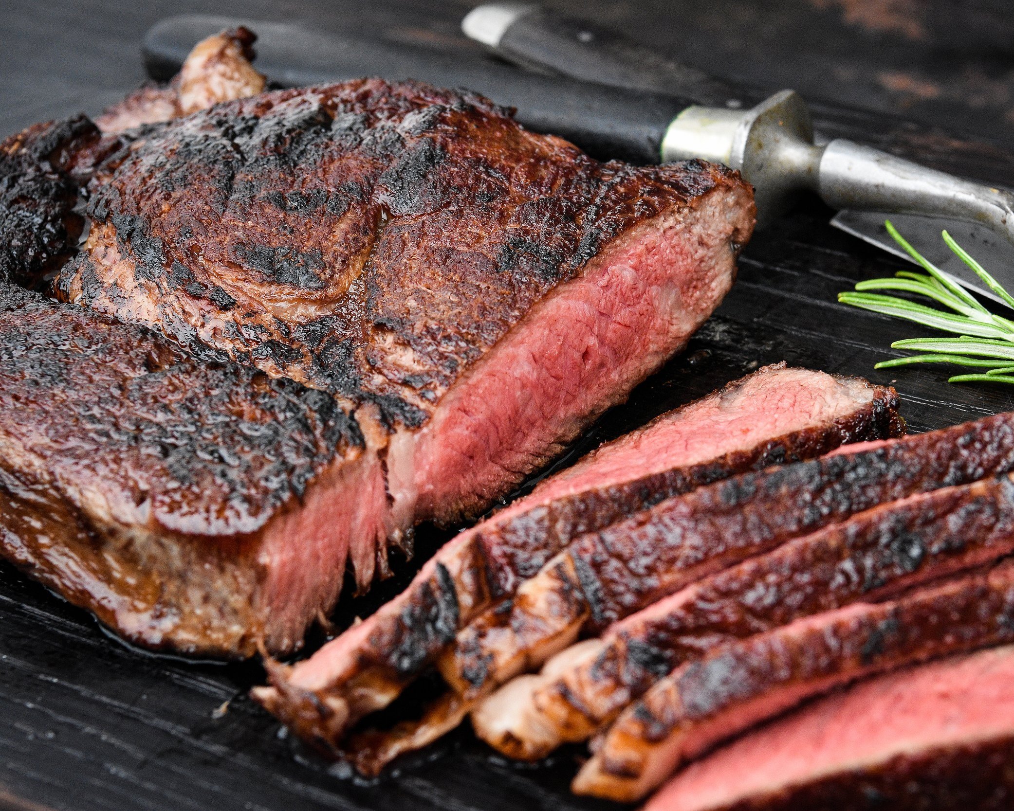 beef_cooked_ribeye Juicy, perfectly cooked steak with a charred exterior and pink, tender interior, sliced and served on a wooden cutting board alongside a knife and a sprig of rosemary. Ideal for a gourmet meal or grilling recipes.