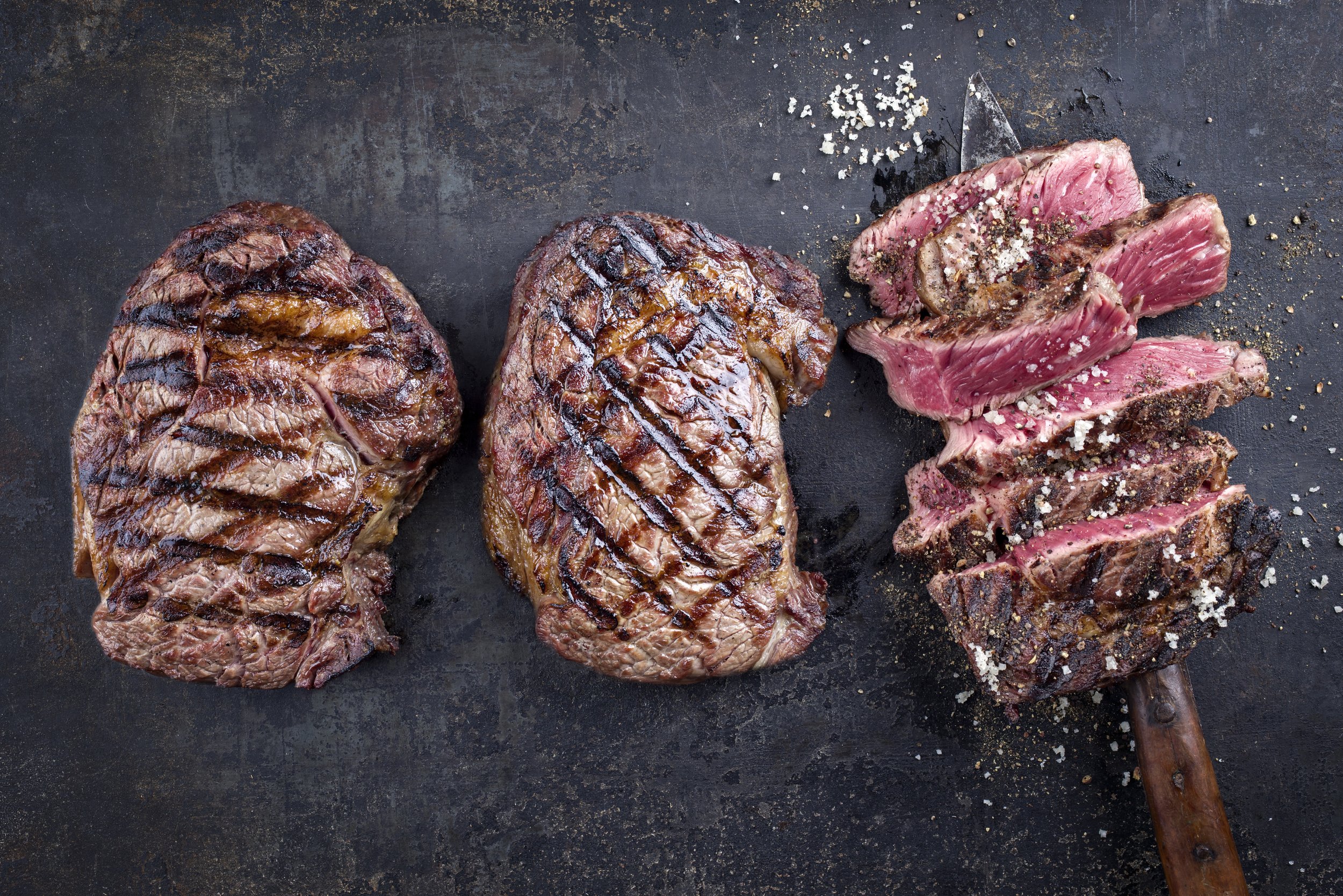 Tree Barbecue Wagyu Entrecote Steaks as top view on old Metall Sheet Grilled beef steaks with distinctive grill marks, showcasing a juicy texture, served alongside sliced portions seasoned with coarse salt and spices on a dark rustic background.