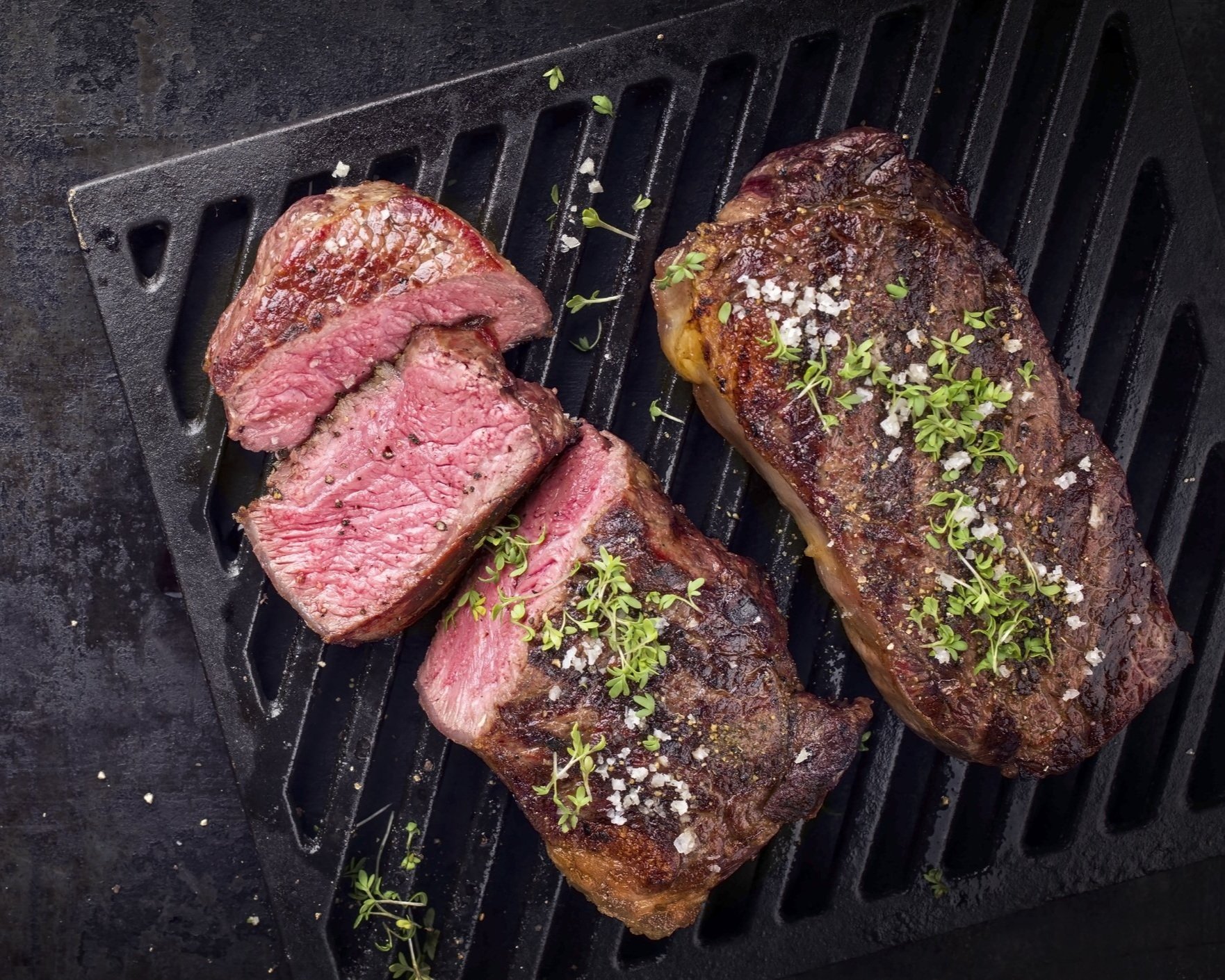 Juicy grilled steak slices garnished with fresh herbs and sea salt, displayed on a black grill plate, showcasing a perfectly cooked medium-rare interior.