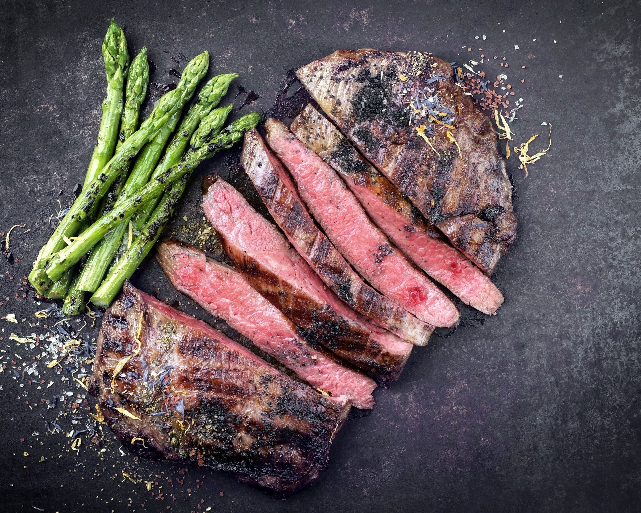 beef_flatironsteak_cooked_sliced Grilled steak slices served with roasted asparagus, garnished with herbs and spices on a dark background. Perfect for a delicious and healthy meal.