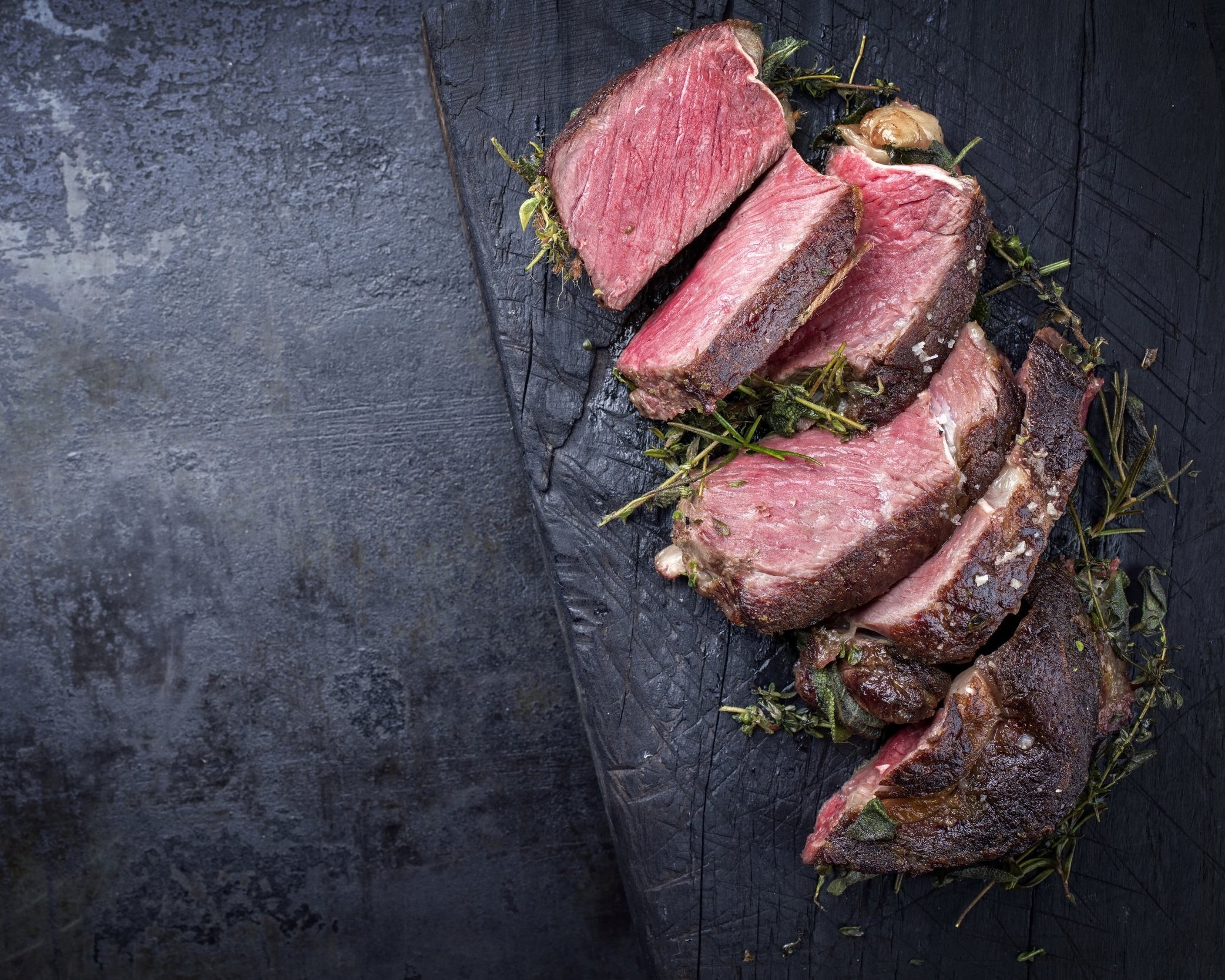 Sliced medium-rare beef resting on a dark wooden cutting board, garnished with herbs, showcasing juicy texture and rich color. Perfect for gourmet meal presentations or barbecue recipes.