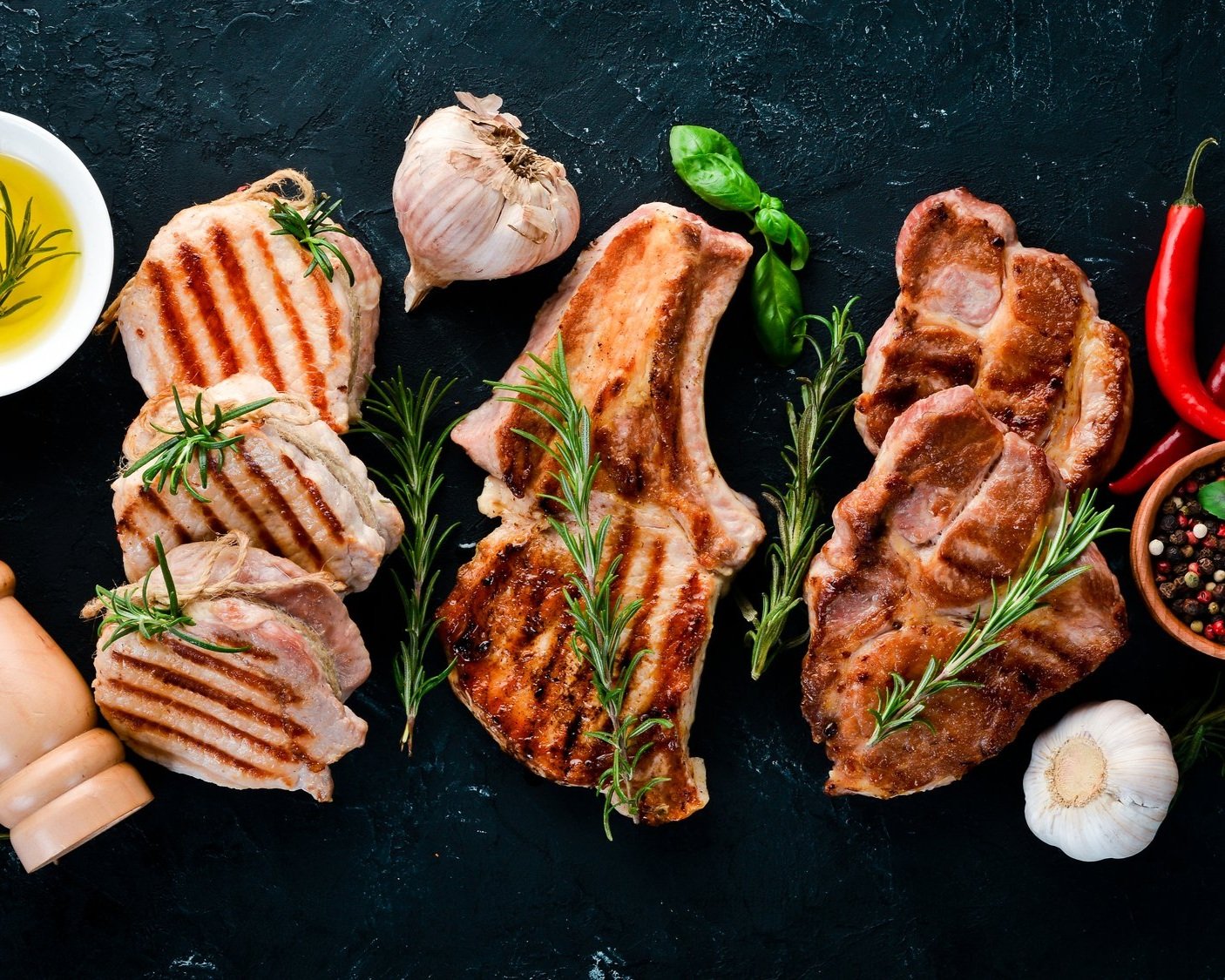 pork_cuts_cooked_tenderloin_chops Grilled pork chops garnished with fresh rosemary, garlic, and basil, accompanied by olive oil and colorful chili peppers on a dark textured background. Perfect for a delicious meal or barbecue inspiration.
